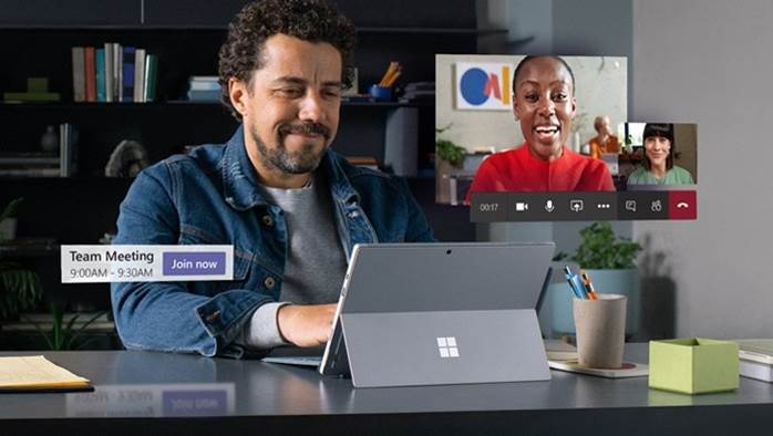 Four people having a Teams meeting in a conference room with five other people joining via video, visible on a large screen at the front of the room. The Teams “join” button floats over the photograph